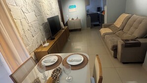 Een moderne woonkamer met een eethoek, met een wand met een stenen textuur, een flatscreen televisie, een bank en een keuken op de achtergrond.