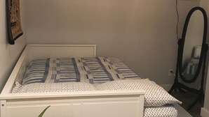 A small, single-bed room with a wooden headboard, a bedside table with a plant, a window with a view of the outside, and a ceiling fan. - Charming 1-bedroom apartment with AC in peaceful Nyack (Nyack)
