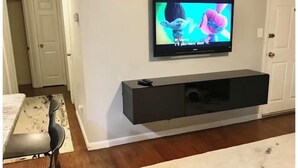 Ein Wohnzimmer mit flachbildfernseher an einer schwarzen Unterhaltungsecke, ein Essbereich mit Tisch und Stühlen sowie ein Flur mit einer Tür.