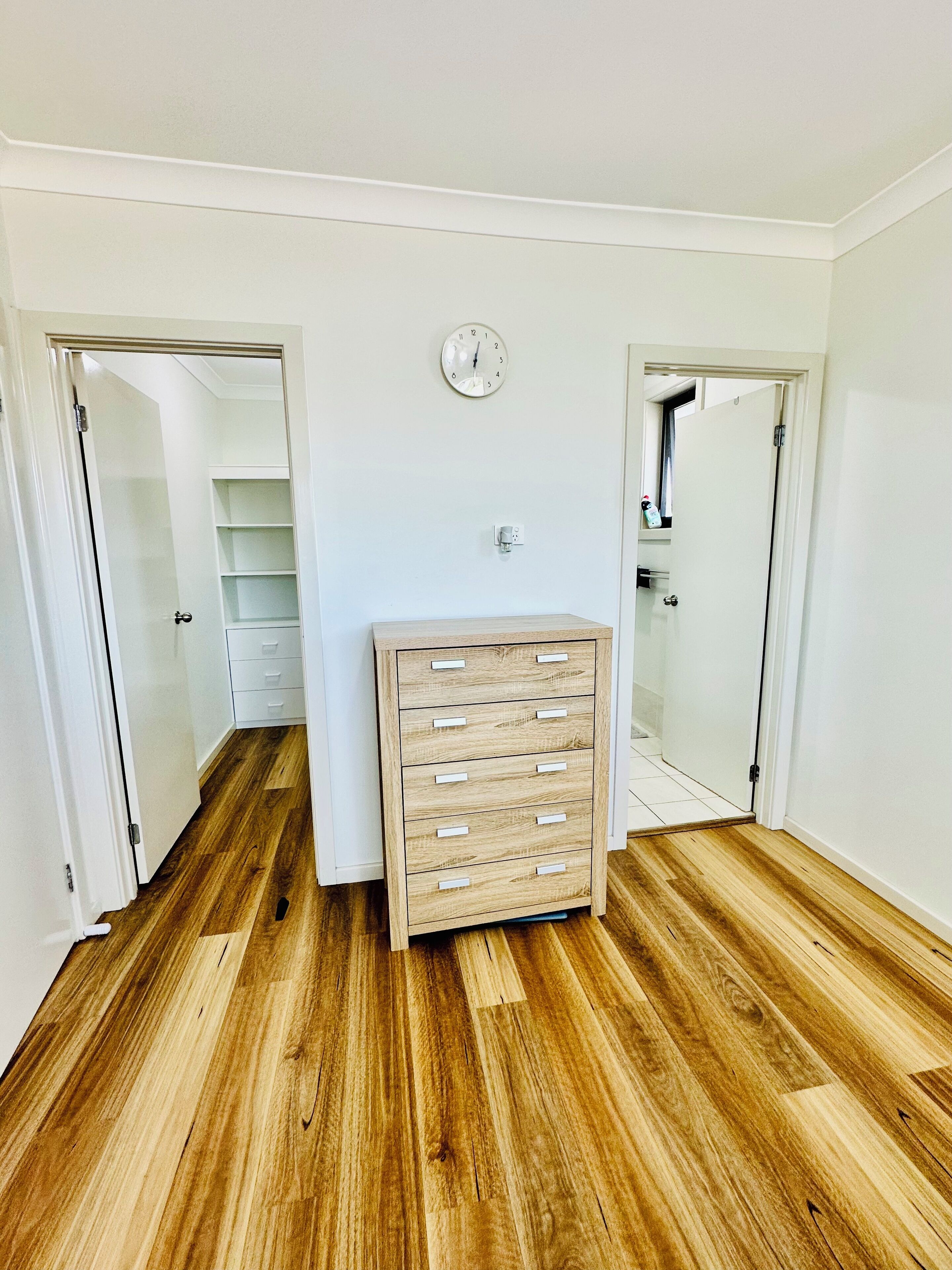 Ein Zimmer mit Holzboden, einer eingebauten Holzkombination mit mehreren Schubladen, einer weißen Uhr an der Wand und einem Kleiderschrank mit Schiebetüren.