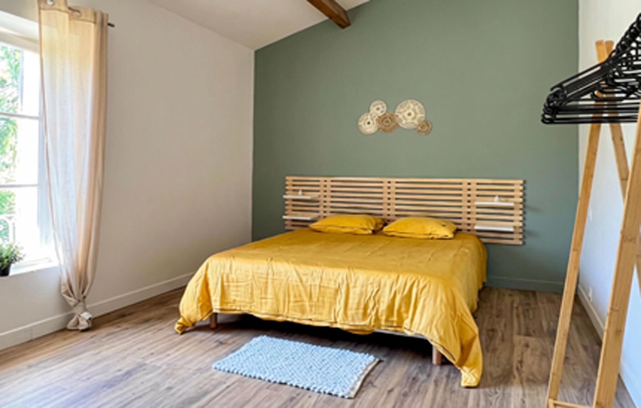 Une chambre à coucher avec un cadre de lit en bois, une literie jaune, un tapis bleu et une fenêtre avec des rideaux.