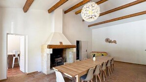 Un'ampia sala da pranzo con un lungo tavolo in legno e sedie bianche, un rustico camino e travi a vista al soffitto.