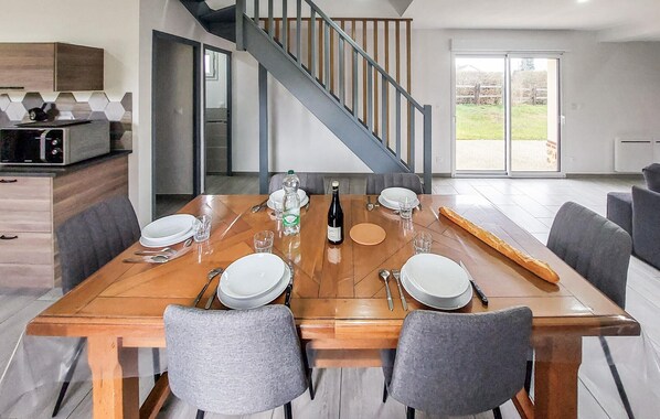 Un coin repas avec une table en bois dressée pour quatre personnes, entourée de chaises grises. Une bouteille de vin et des verres sont posés sur la table. On aperçoit, au fond, un escalier avec une rampe en bois.