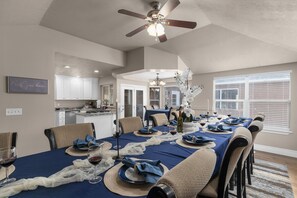 A dining area with a long table set for a meal, a ceiling fan, and a kitchen in the background.