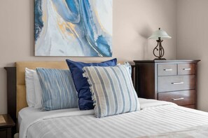 A hotel room with a bed, pillows, a nightstand, a lamp, and a dresser.