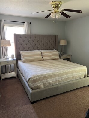 A bedroom with a tufted headboard, a bed with striped bedding, a nightstand with a lamp, and a ceiling fan.