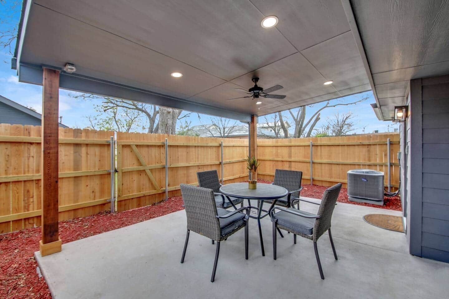 An outdoor patio with a round table and chairs, a ceiling fan, and a wooden fence in the background.