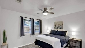 Ein Schlafzimmer mit Bett, Deckenventilator, Nachttisch mit Lampe und einem Fenster mit Vorhängen.