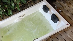 A person sitting by a spa tub with a wooden deck and a small wooden stool with a bottle. - Chalet with hot tub: water directly from the Bridal Veil Waterfall heated to 40°C. (Santana do Riacho)