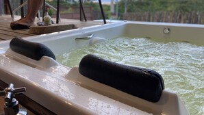 A hot tub with two black bath mats, a wooden deck, and a person sitting on a chair. - Chalet with hot tub: water directly from the Bridal Veil Waterfall heated to 40°C. (Santana do Riacho)