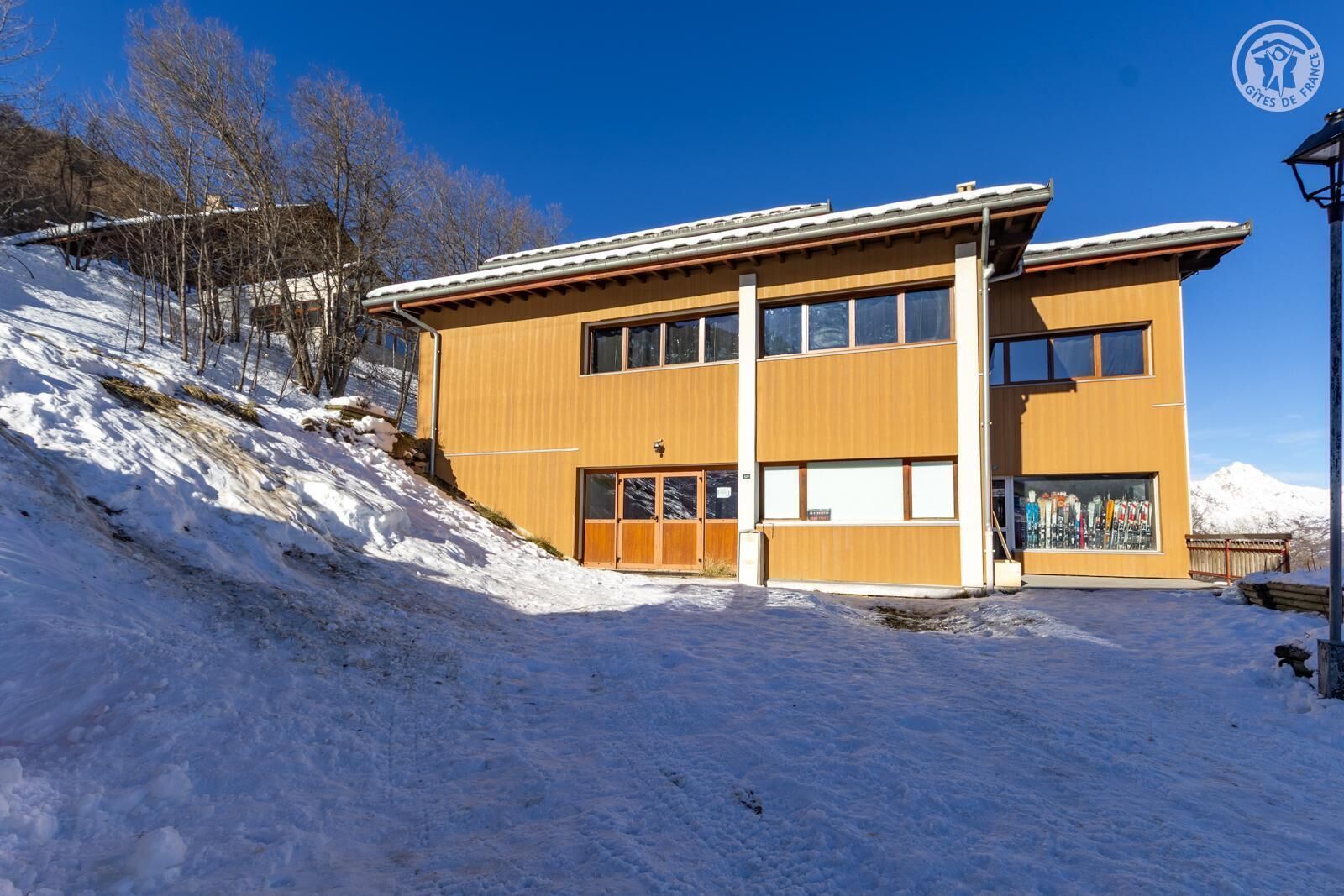 Ein modernes Gebäude mit großen Fenstern und einer Holzfassade, gelegen an einem verschneiten Hang unter blauem Himmel.