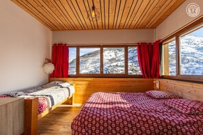 Une pièce chaleureuse avec un plafond en bois, deux lits dotés de draps à motifs floraux et de grandes fenêtres offrant une vue imprenable sur les montagnes enneigées.