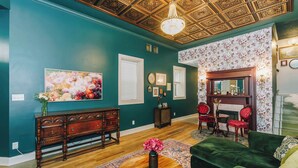 Een kamer met blauwgroene muren, een houten vloer, een groene bank, een ronde salontafel met een vaas, een bloemschilderij en een kroonluchter.