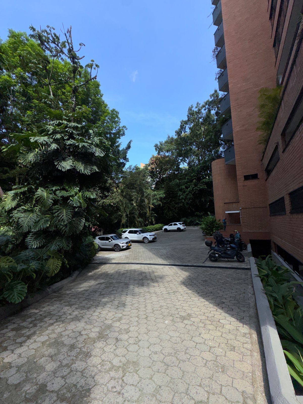 A paved courtyard with parked cars and motorcycles, surrounded by lush greenery and a brick building.