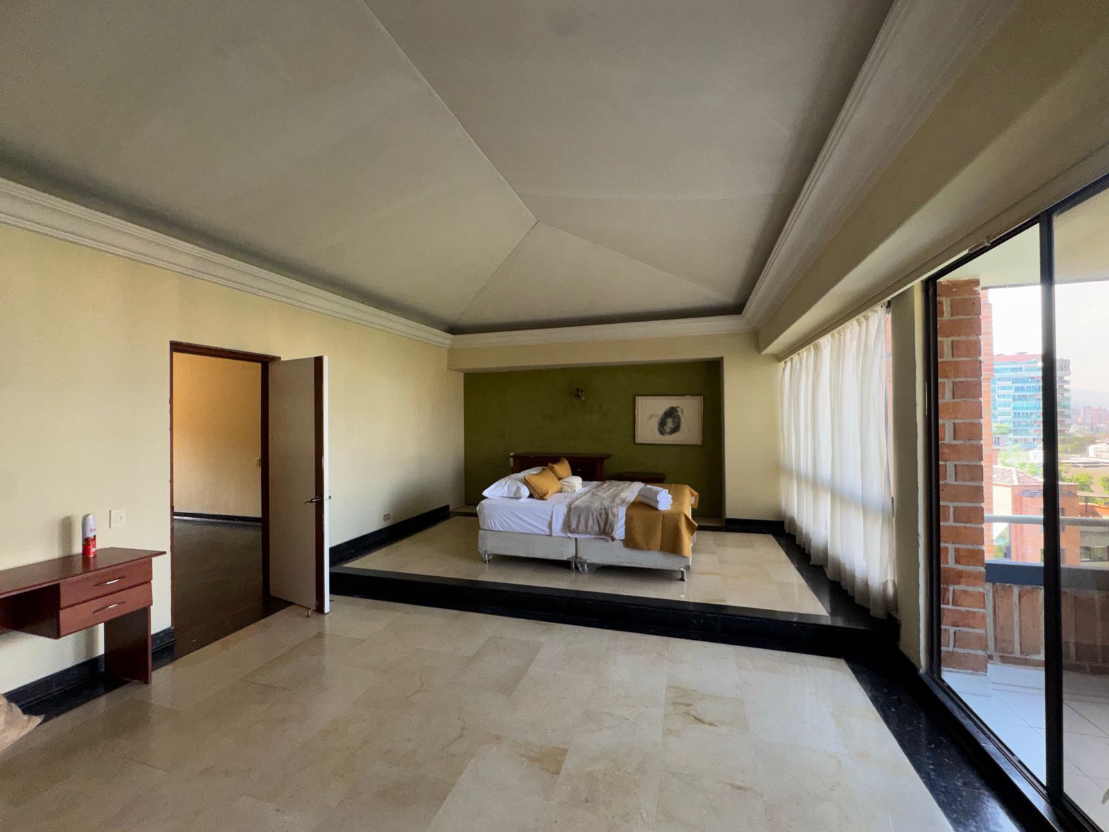 A spacious bedroom with a large bed, a desk, and a view of the city through a large window.