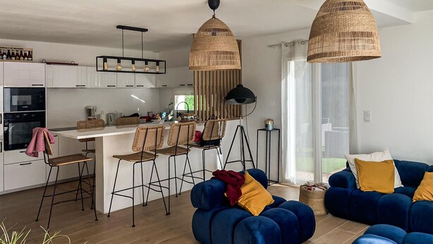 Eine moderne Küche mit weißen Schränken, einer zentralen Kochinsel mit Barhocker und einem Essbereich mit blauem Sofa und Kissen.