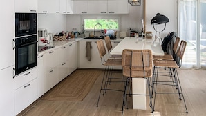 Eine moderne Küche mit weißen Schränken, einem eingebauten Backofen, einem Essbereich mit Korbstühlen und einem großen Fenster für natürliches Licht.