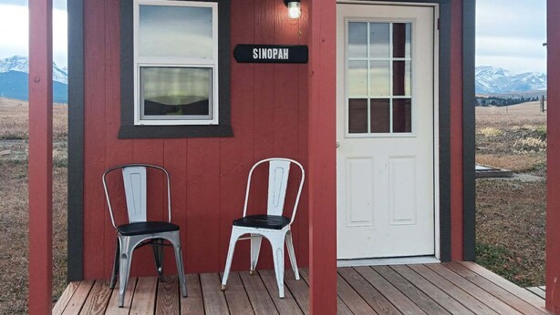 A small wooden cabin with a red exterior, a white door, and two white chairs on the porch. A sign above the door reads "SINOPAH." - Sinopah #5 (East Glacier Park)