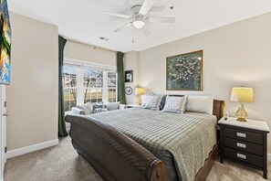 Une chambre à coucher avec un lit, une table de chevet, un ventilateur de plafond, une fenêtre avec des rideaux et un tableau accroché au mur.