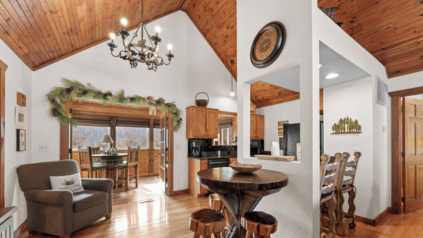 A spacious living area with a wooden ceiling, a chandelier, a round wooden table with stools, and a cozy armchair.