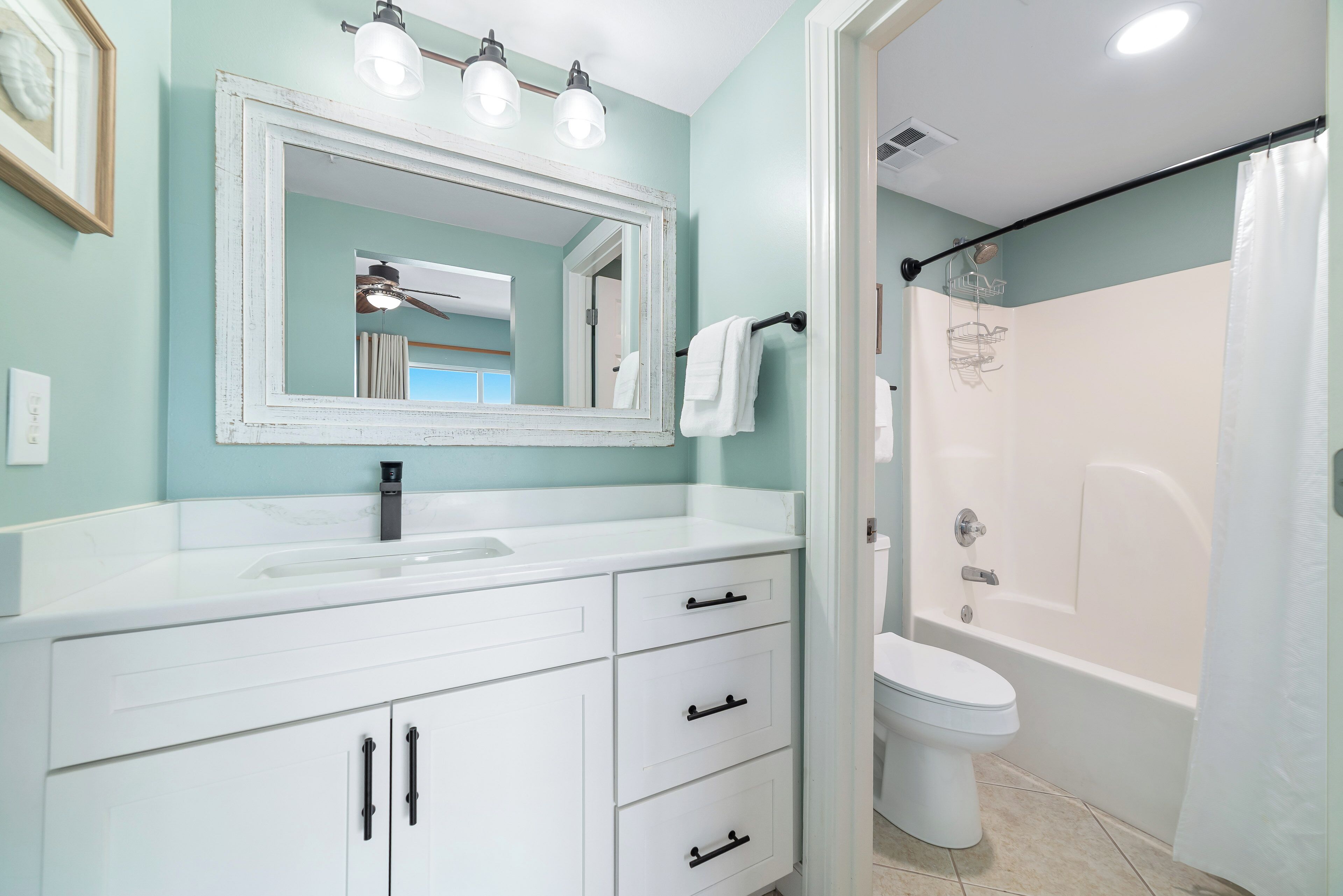 Une salle de bain avec un meuble-lavabo blanc, un grand miroir, un ventilateur de plafond, des toilettes et un espace douche.