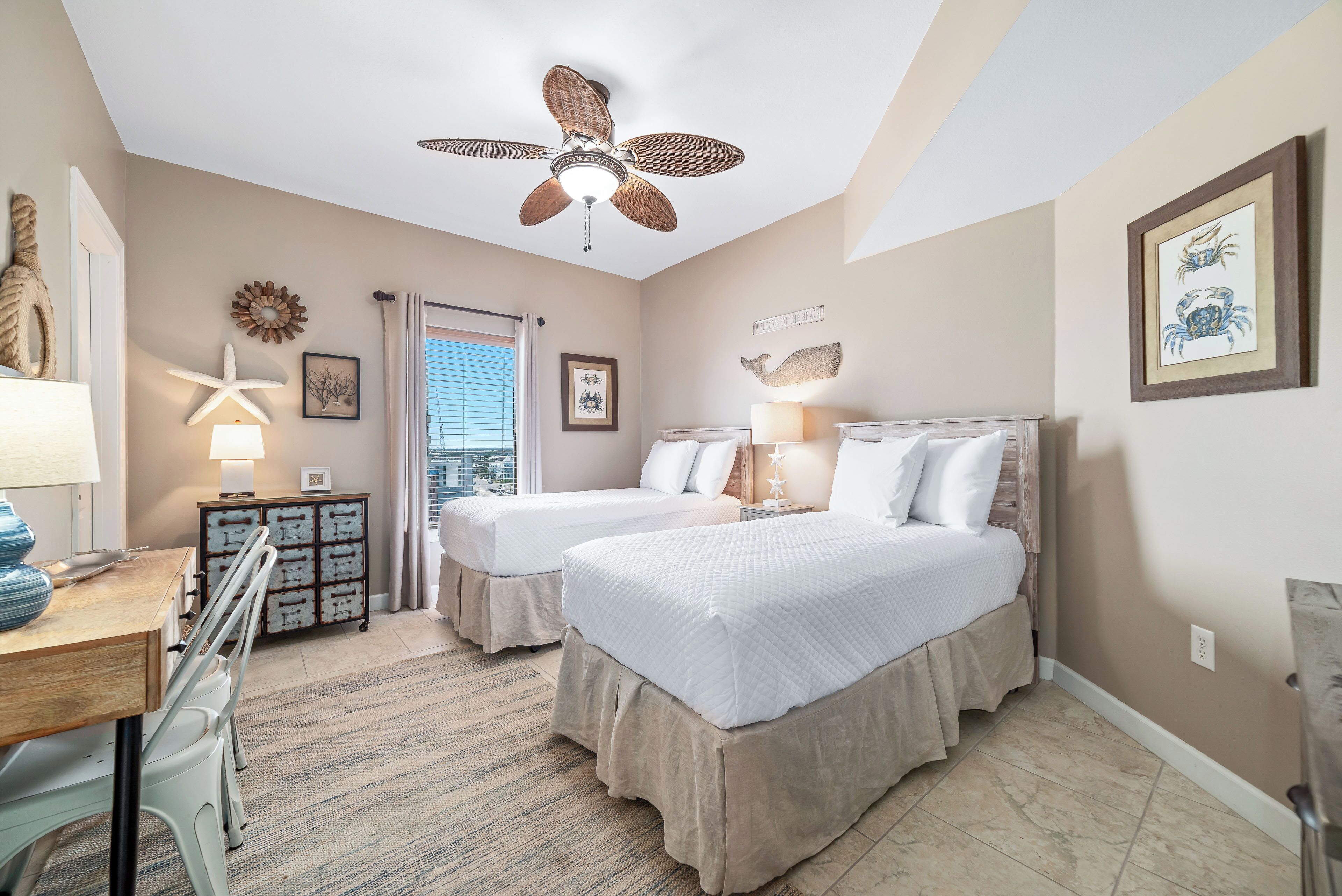 A bedroom with two beds, a ceiling fan, a desk, and a view of the city.