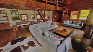 Ein großzügiges Wohnzimmer mit einer großen Eckcouch, einem Couchtisch und einem Kuhfellteppich. Eine Treppe führt zu einer oberen Ebene, und der Raum hat Holzwände und -böden.