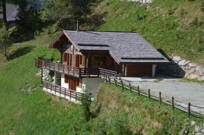 Una cabaña de madera con balcón, situada en una ladera con un sendero de grava y una valla de madera.