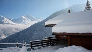 Eine Holzhütte mit schneebedecktem Dach, umgeben von einem Holzzaun und verschneitem Gelände, mit einer Bergkulisse im Hintergrund.