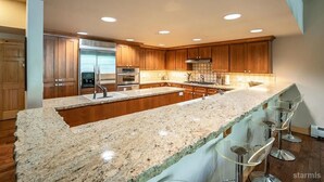 Une cuisine moderne dotée de plans de travail en granit, d’appareils électroménagers en acier inoxydable, d’armoires en bois et d’un îlot central avec des tabourets de bar.