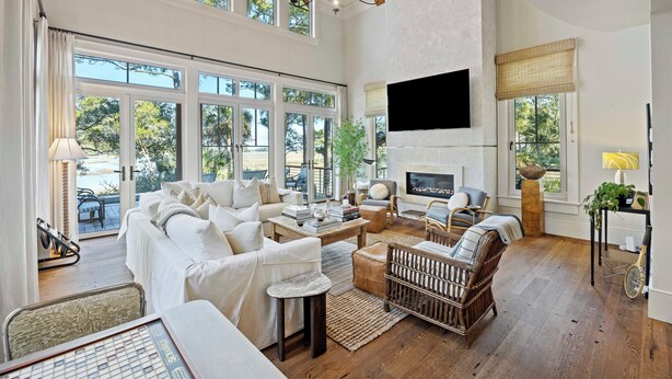 A spacious living room with a large sofa, a coffee table, and a fireplace. There are multiple windows providing natural light and a view of the outdoors. - Stunning Marsh Views! Plunge Pool, Quiet Kiawah 3BR Home! (Kiawah Island)