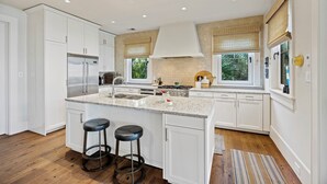 A modern kitchen with white cabinets, a stainless steel refrigerator, a large island with bar stools, and a window with a view of greenery. - Stunning Marsh Views! Plunge Pool, Quiet Kiawah 3BR Home! (Kiawah Island)