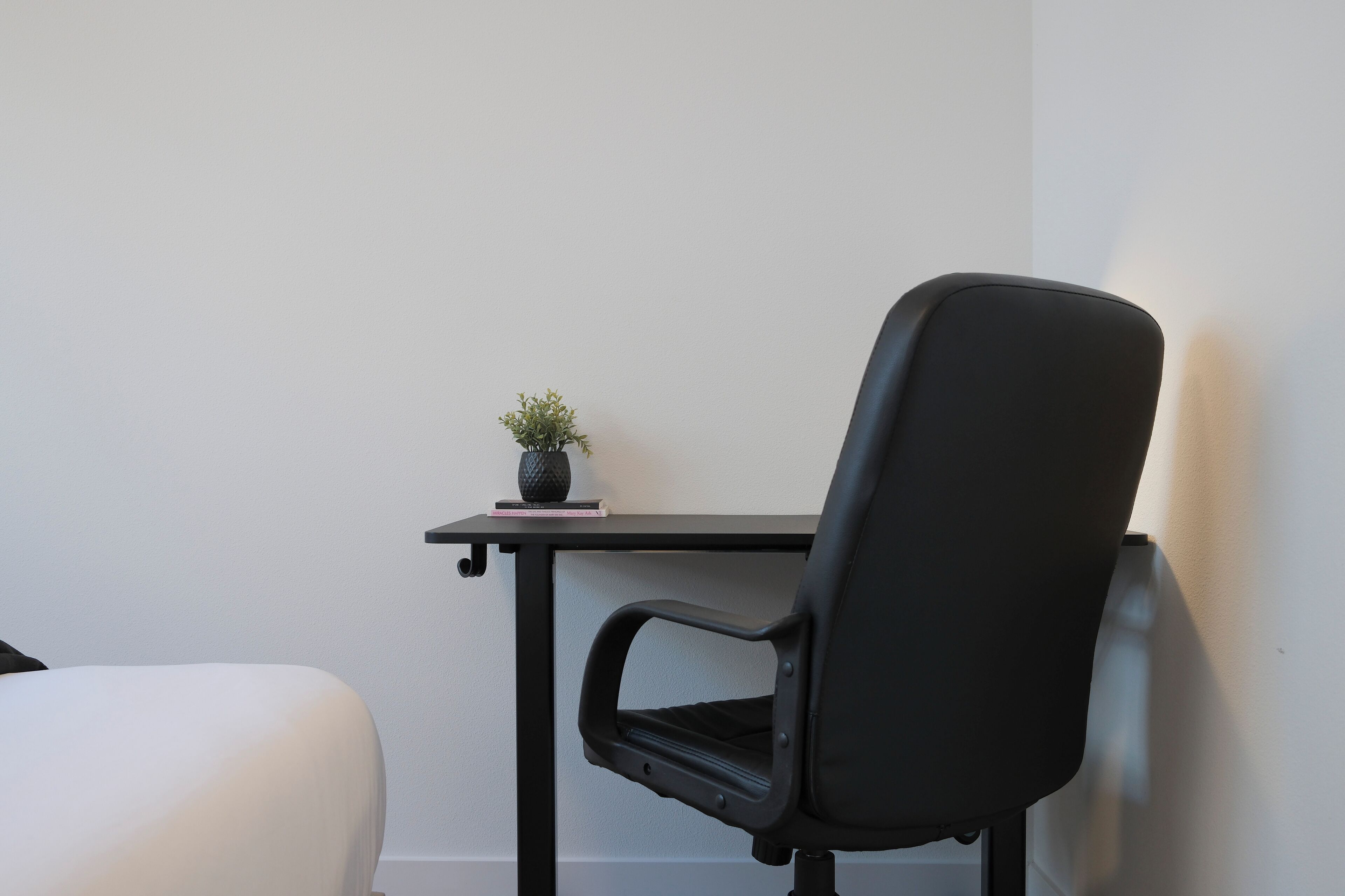 A room with a black office chair, a small potted plant, and a white bed.