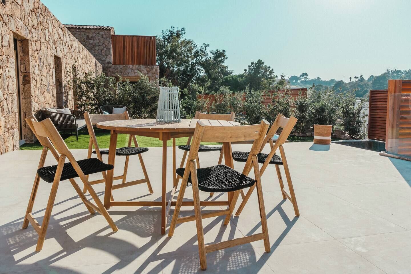Une terrasse extérieure avec une table ronde en bois et six chaises, entourée d’un mur de pierre et de verdure.