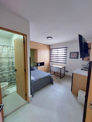 Quarto compacto com cama, escrivaninha, chuveiro e banheiro pequeno.