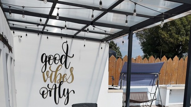 Eine Außenpatio mit Korbmöbeln, einem Glastisch und einer Wand mit dem Schriftzug 'good vibes only'.