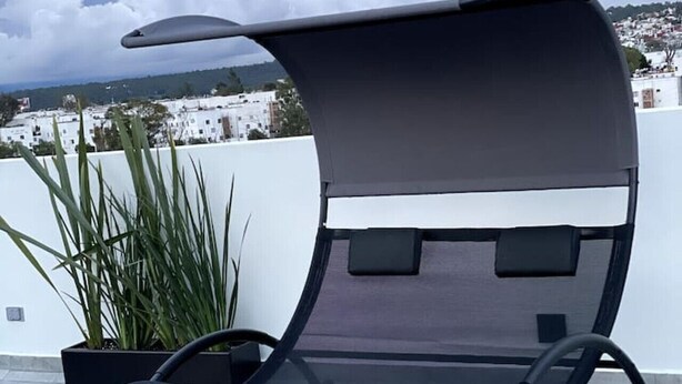 Un área de estar moderna al aire libre con un banco acolchado y una marquesina, rodeada de plantas en macetas y vistas a un paisaje urbano.