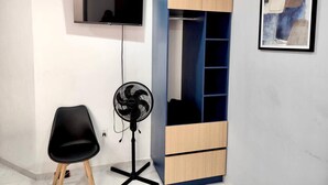 Una stanza con una TV montata a muro, una sedia nera, una ventola portatile e un armadio a muro con ripiani.