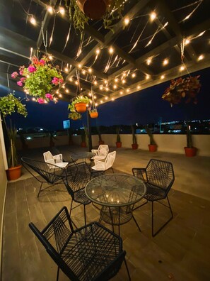 Een terras buiten met hangplanten, lichtslingers en zitplaatsen.
