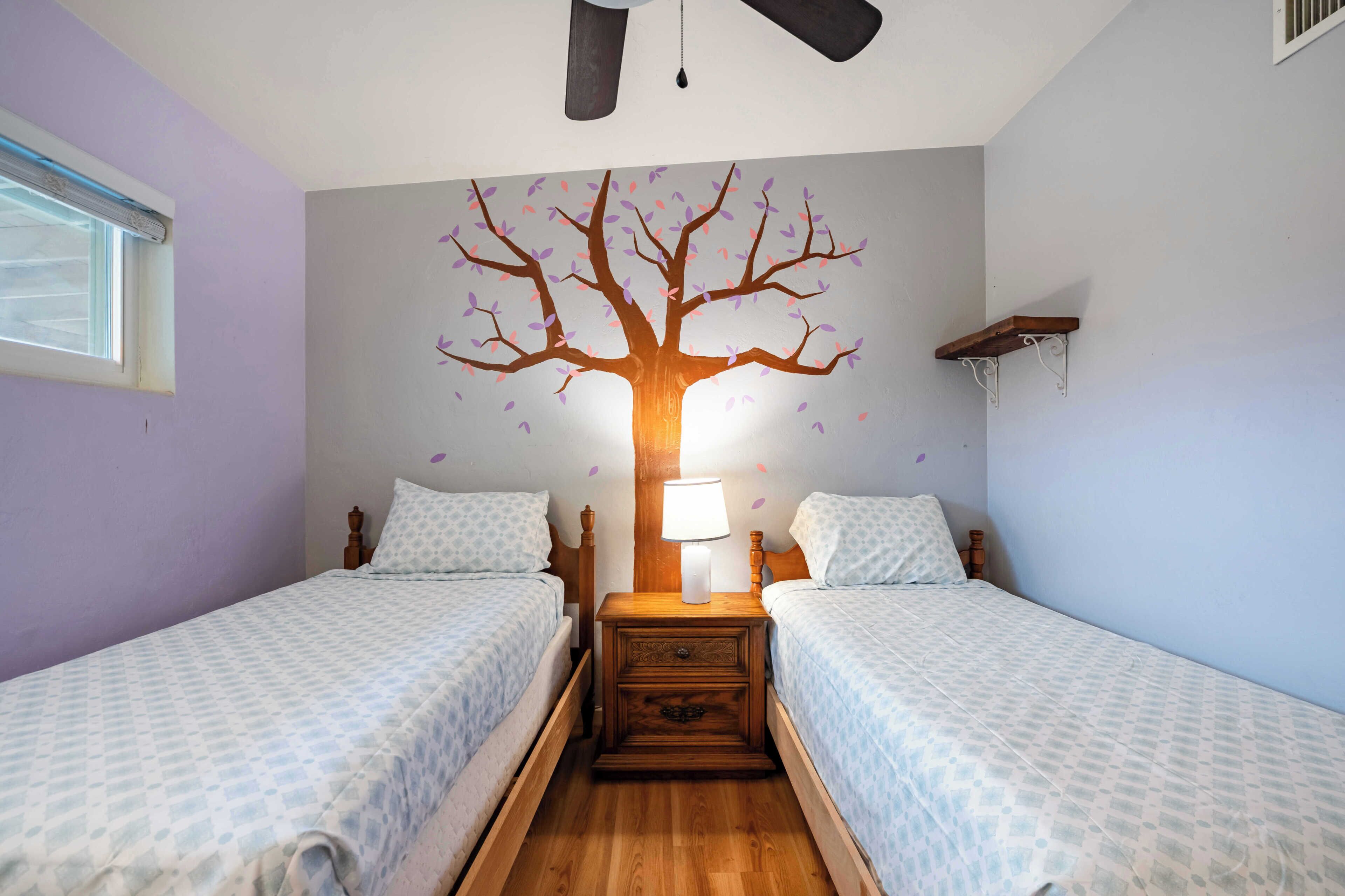 A bedroom with two beds, a nightstand with a lamp, a wall mural of a tree, and a window.