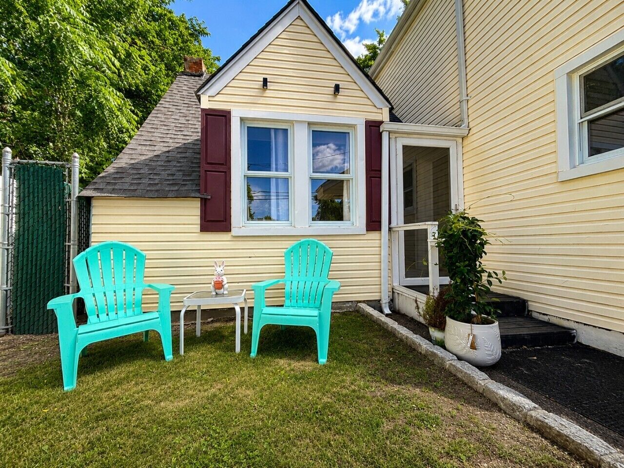 A house with a small front yard, two turquoise Adirondack chairs, a white table, and a potted plant.