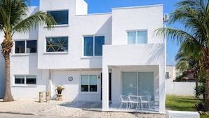 Un edifici modern i blanc amb nombroses finestres, una zona d'estar exterior coberta i palmeres.