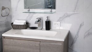 Un bany amb una paret revestida de marbre, un lavabo blanc amb un moble de fusta a sota, una prestatgeria de vidre sobre el lavabo, i un mirall.