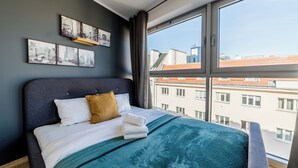 A modern hotel room with a large bed, a view of the city through a large window, and cityscape artwork on the wall.