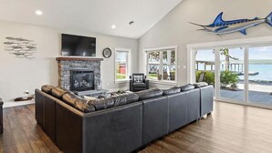 A modern living room with a stone fireplace, a large flat-screen TV, and a view of the ocean through large windows. - Mott's Lakefront Landing: Dog-Friendly Lakefront Escape on Seneca Lake! (Lodi)