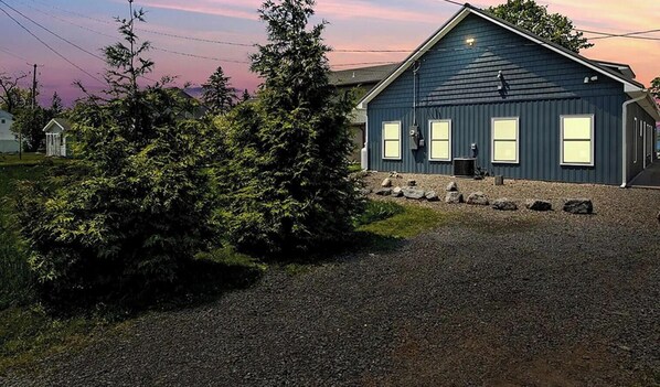 A two-story building with a dark blue exterior, a gravel driveway, and a small garden area with a tree. - Mott's Lakefront Landing: Dog-Friendly Lakefront Escape on Seneca Lake! (Lodi)