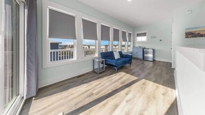 A modern living room with a blue sofa, large windows, and a kitchen area. - Sandy Bottoms by Carolina Getaway: Elevator Home w/ Ocean Views & Steps to Beach (Surf City)