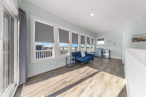 A modern living room with a blue sofa, large windows, and a kitchen area. - Sandy Bottoms by Carolina Getaway: Elevator Home w/ Ocean Views & Steps to Beach (Surf City)