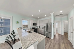 A modern kitchen with granite countertops, stainless steel appliances, and a dining area with chairs. - Sandy Bottoms by Carolina Getaway: Elevator Home w/ Ocean Views & Steps to Beach (Surf City)