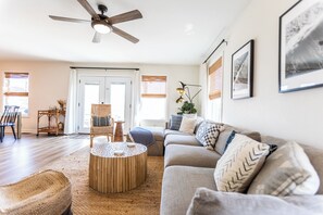 Ein Wohnzimmer mit Deckenventilator, einer Couch, einem Couchtisch, einem Korbstuhl und einem Essbereich mit Tisch und Stühlen.